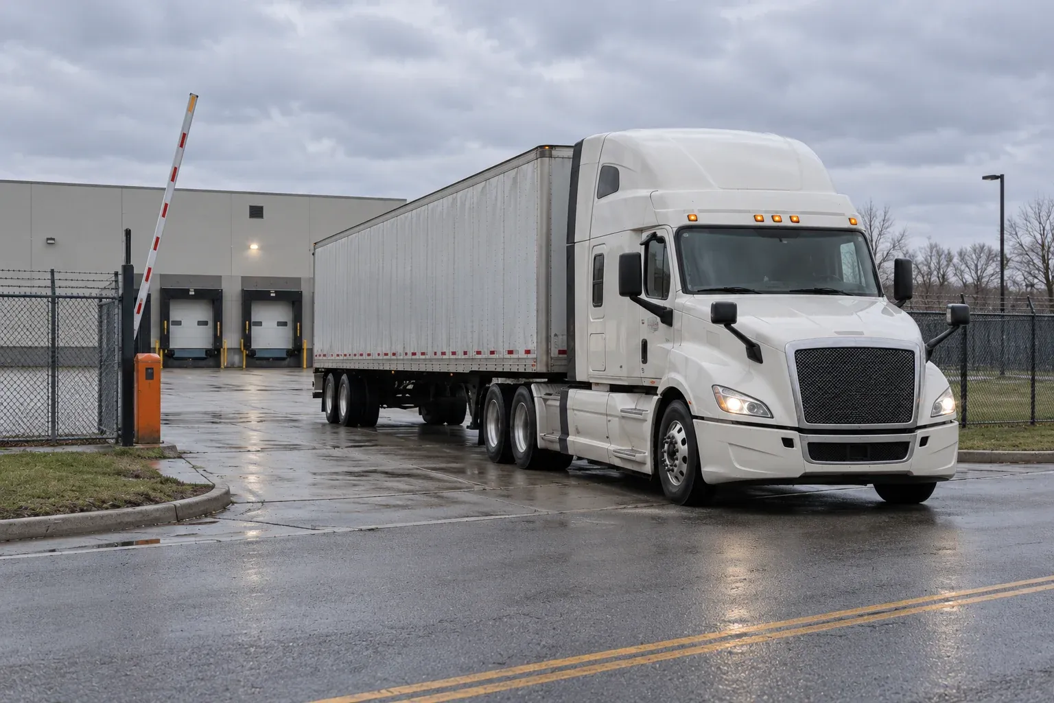 An ivory tractor-trailer crossing an open exit gate toward the road, with two warehouse dock doors behind it.
